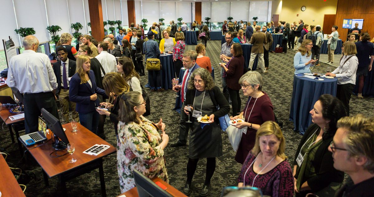 A BUSDC poster session with lots of people talking , eating, and presenting posters in a large room filled with physical and digital posters.